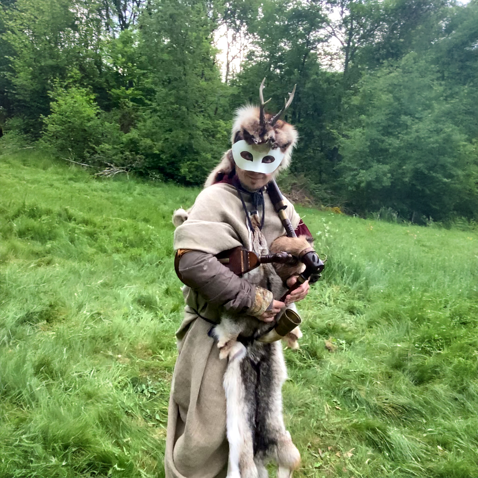 Person in medieval costume standing in a lush green meadow. The person is wearing a white mask with antlers and fur, and is holding a traditional musical instrument. The costume includes a beige cloak and fur accessories, giving a historical and nature-inspired look. The background is a dense forest with vibrant greenery.